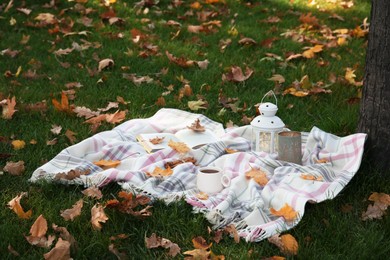 Photo of Plaid, cup of coffee, book, lantern and candle on green grass covered with autumn leaves