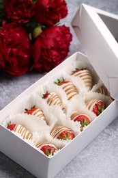 Photo of Delicious chocolate covered strawberries in box and flowers on light grey table