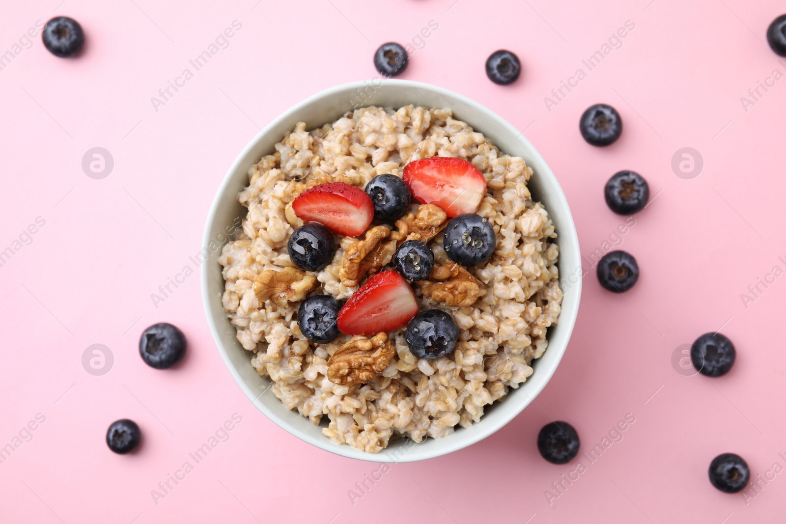 Photo of Tasty oatmeal with strawberries, blueberries and walnuts in bowl surrounded by fresh berries on pink background, flat lay