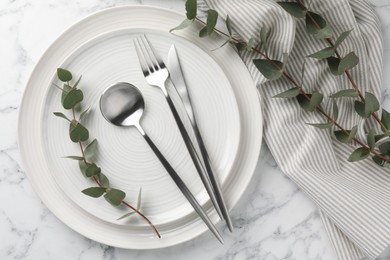 Photo of Stylish setting with cutlery, napkin, eucalyptus branches and plates on white marble table, top view