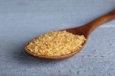 Photo of Spoon with uncooked bulgur on grey wooden table, closeup