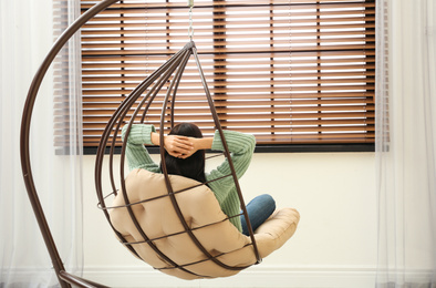 Photo of Young woman relaxing in hanging chair near window at home, back view