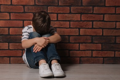 Photo of Upset boy sitting on floor near brick wall, space for text. Children's bullying