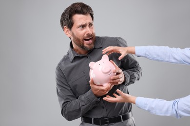 Photo of Scared man trying to protect piggy bank from woman on light grey background. Be careful - fraud