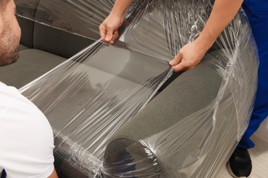 Photo of Workers wrapping sofa in stretch film indoors, closeup