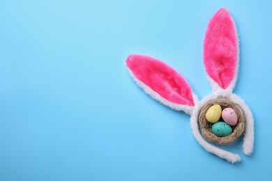 Headband with Easter bunny ears and dyed eggs in nest on light blue background, flat lay. Space for text