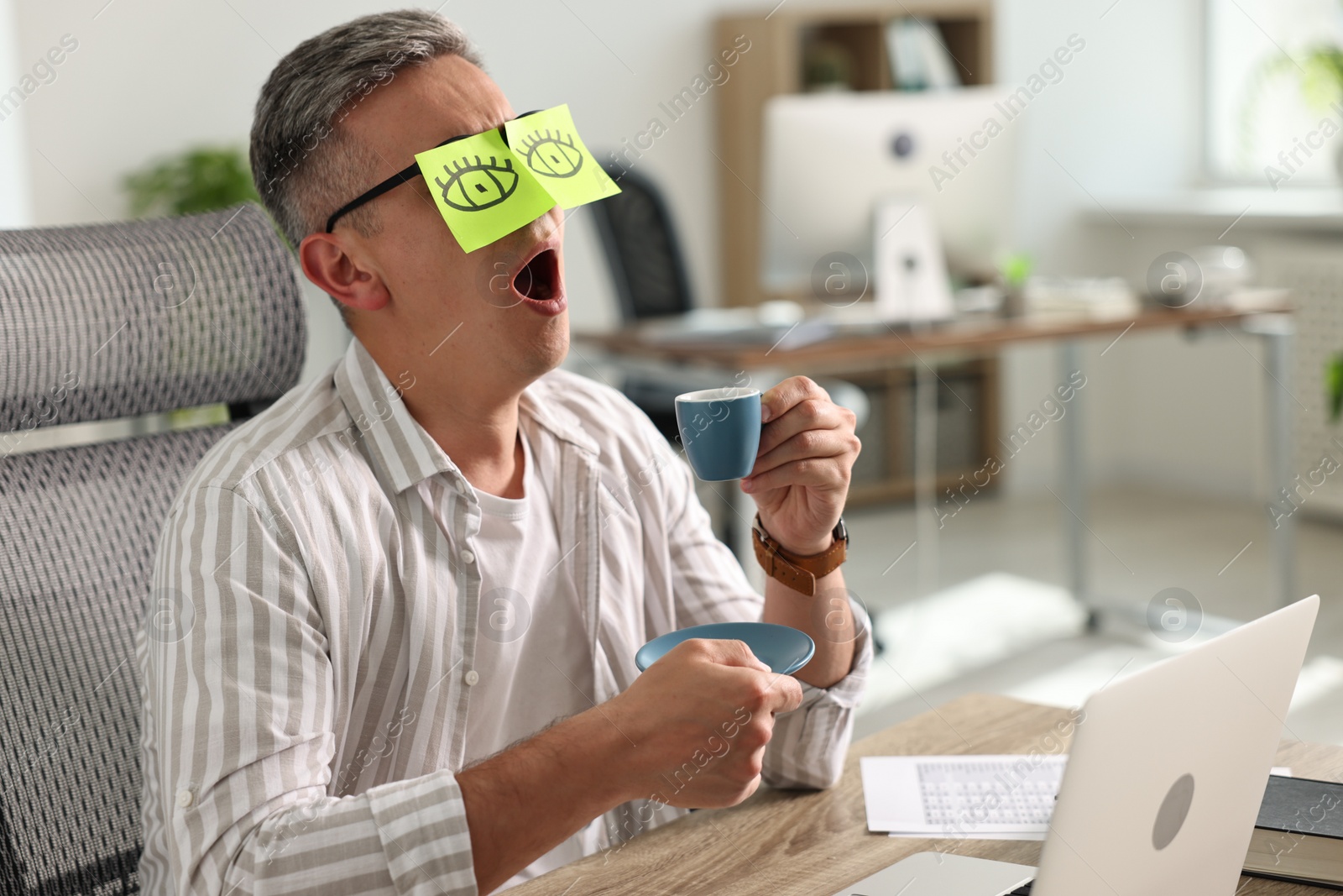 Photo of Man with fake eyes painted on sticky notes holding cup of drink and yawning at workplace in office