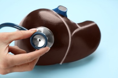 Doctor with stethoscope examining liver model at light blue background, closeup