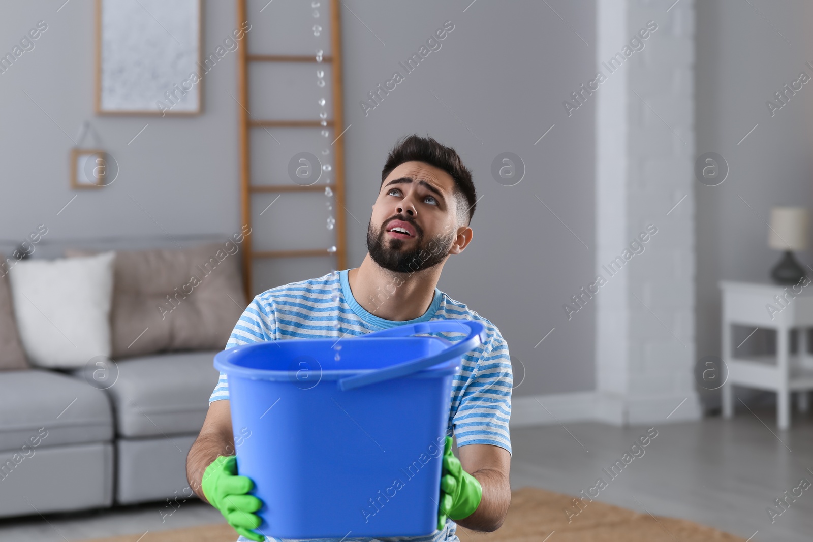 Photo of Young man collecting leaking water from ceiling at home, space for text. Time to call roof repair service