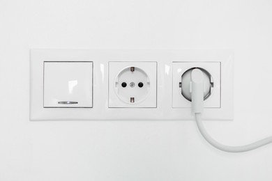 Power sockets with inserted plug and light switch on white wall indoors