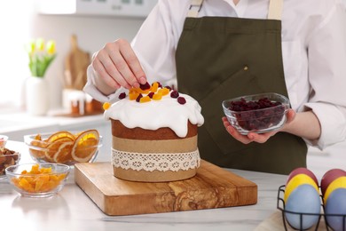 Woman decorating delicious Easter cake with dried cranberries at white marble table in kitchen, closeup