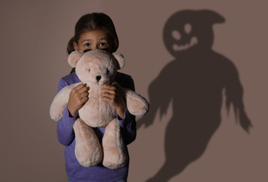 Scared little girl with teddy bear suffering from sciophobia and phantom behind her. Irrational fear of shadows