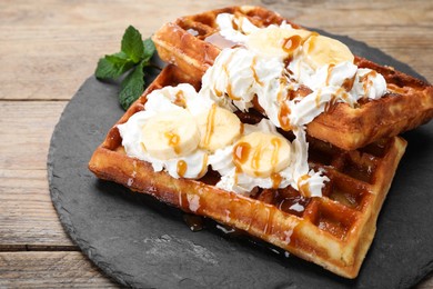 Photo of Slate plate of delicious Belgian waffles with banana, whipped cream and caramel sauce on wooden table, closeup