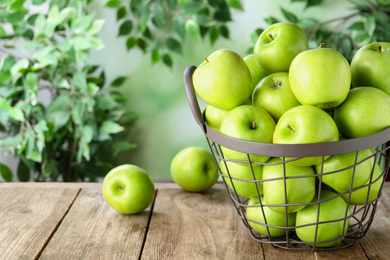 Photo of Ripe green apples in metal basket on wooden table against blurred background. Space for text