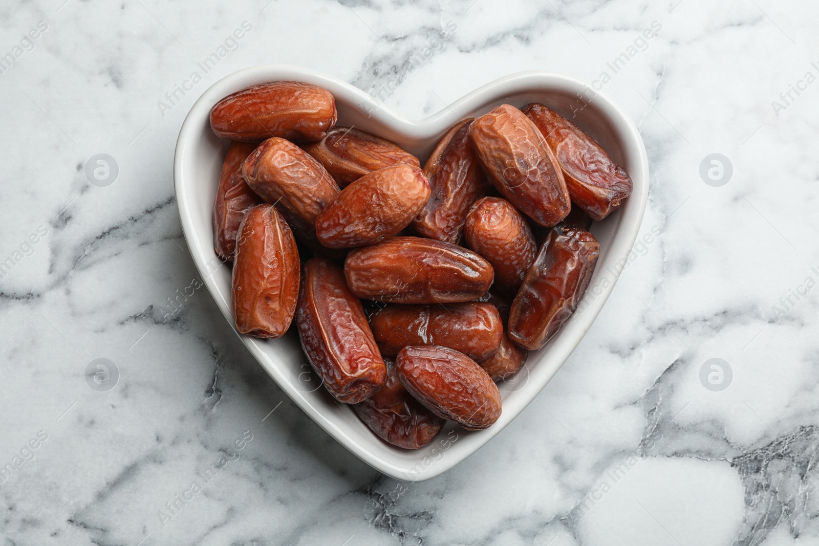 Photo of Heart shaped bowl with sweet dried date fruits on marble background, top view