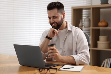 Photo of Young man with cup of coffee watching webinar at table in room