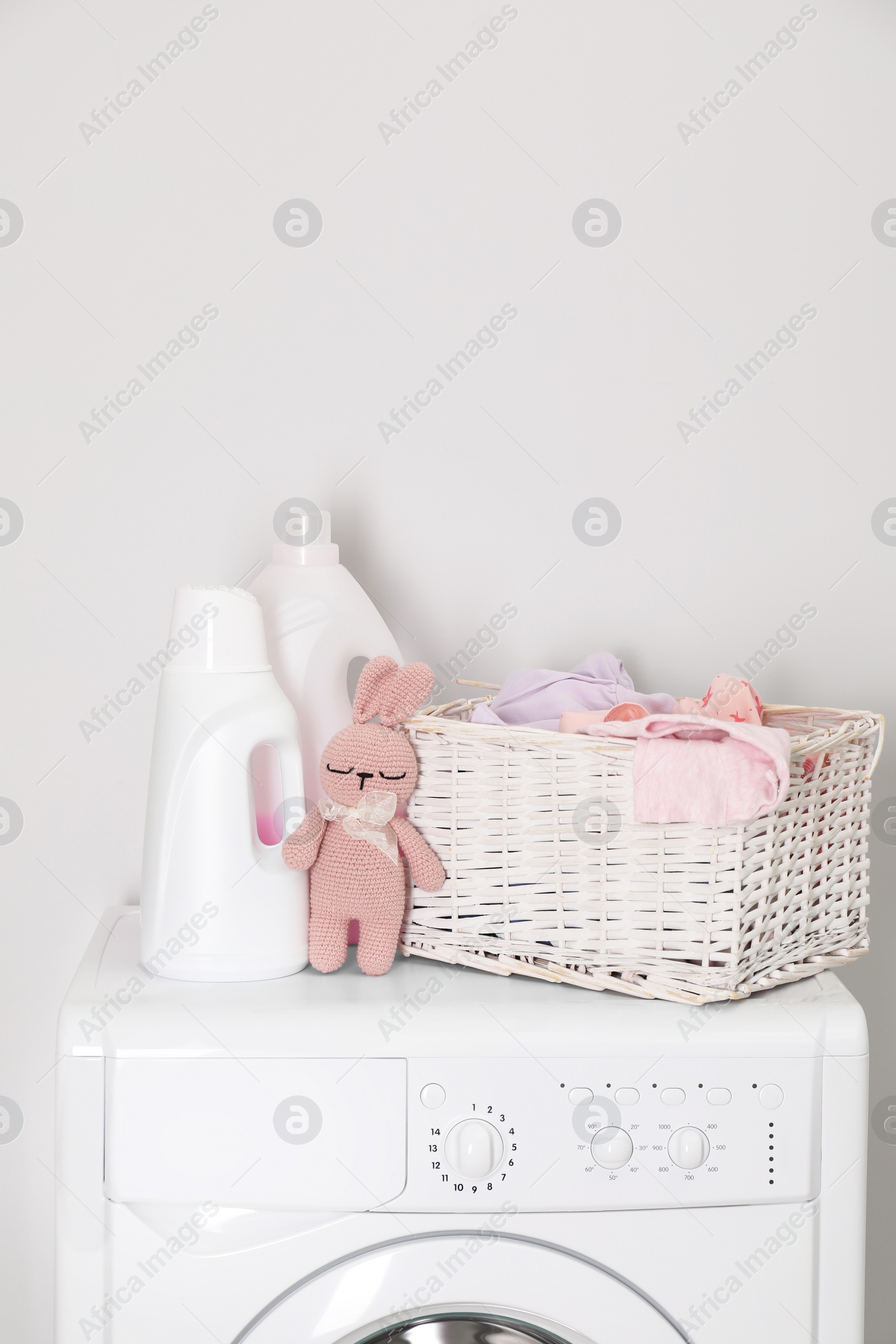 Photo of Baby clothes in wicker basket, laundry detergents and toy bunny on washing machine near light wall, space for text