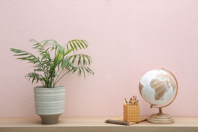 Beautiful plant in pot, globe and stationery on wooden table near pink wall. Interior accessories