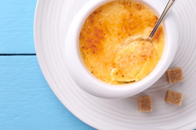 Photo of Delicious creme brulee in bowl, sugar cubes and spoon on light blue wooden table, top view