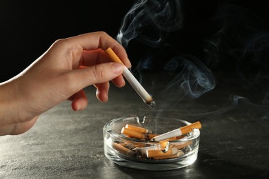 Photo of Woman holding smoldering cigarette over glass ashtray at grey table against black background, closeup