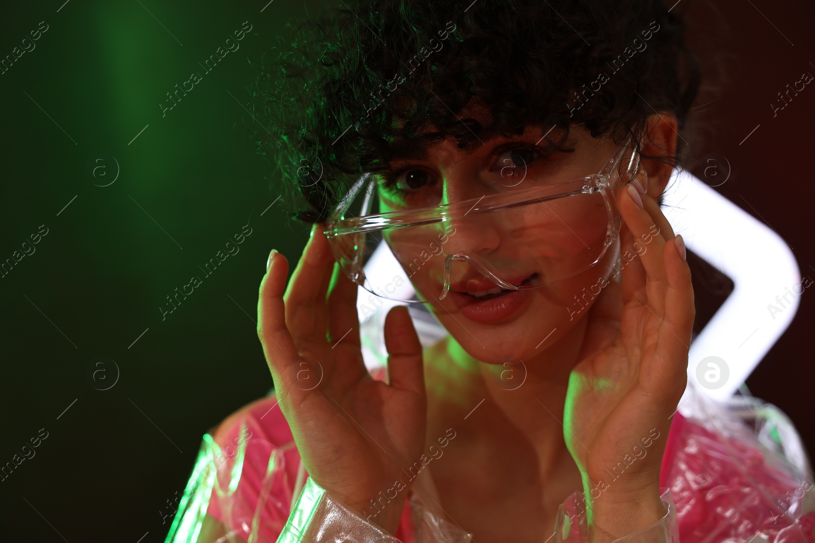 Photo of Beautiful young woman in transparent coat and sunglasses with square lamp on color background in neon lights