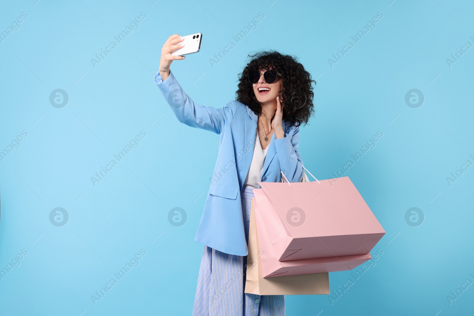 Photo of Happy young woman with shopping bags taking selfie on light blue background