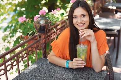 Photo of Young woman with glass of tasty lemonade at table in cafe, outdoors. Natural detox drink