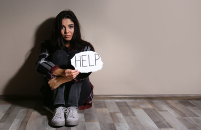 Photo of Abused young woman with sign HELP near beige wall. Domestic violence concept