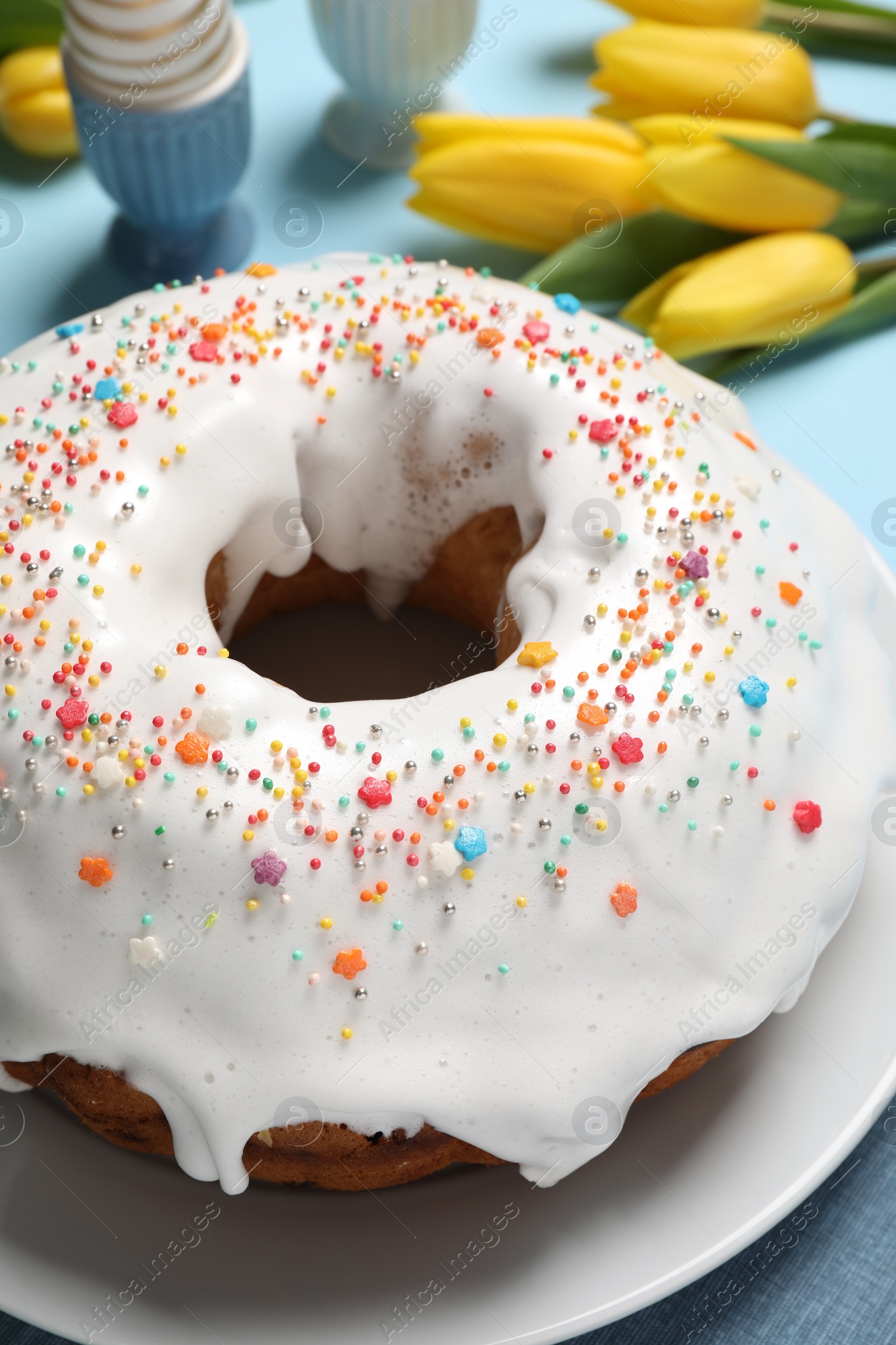 Photo of Delicious Easter cake decorated with sprinkles near beautiful tulips on light blue background, closeup