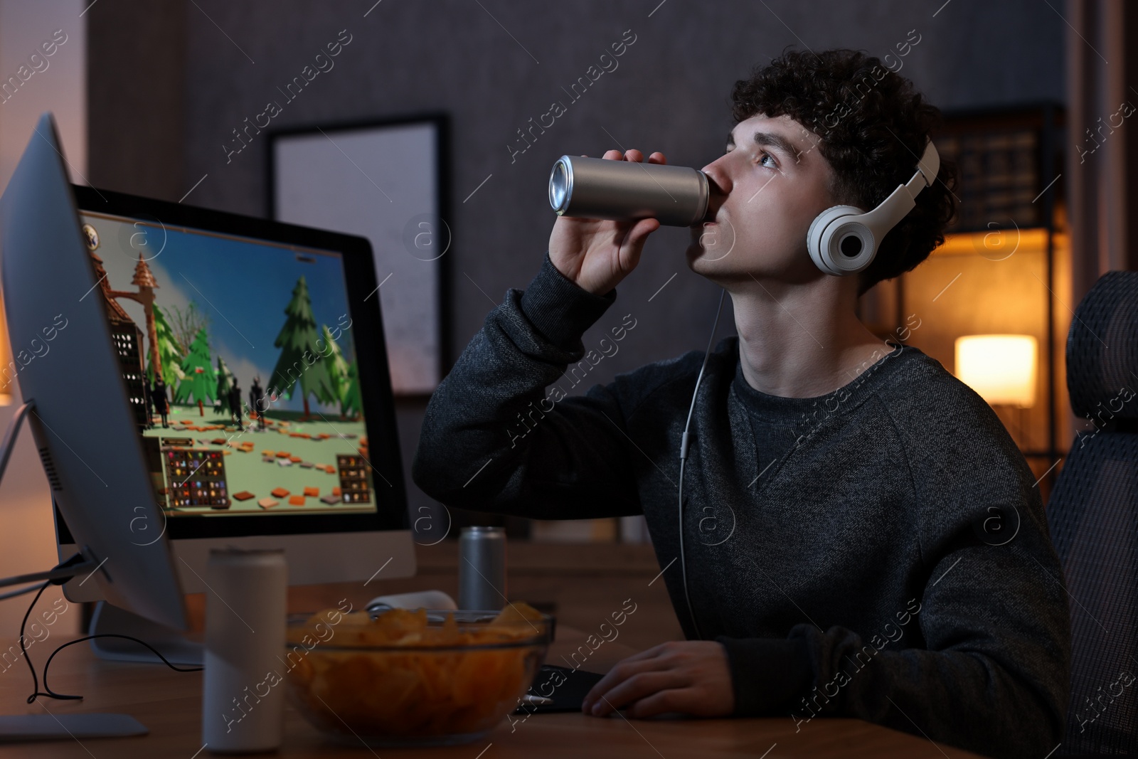 Photo of Young man with energy drink and headphones playing video game at wooden desk indoors