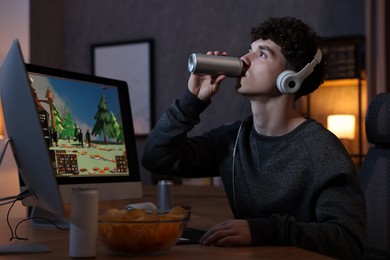 Photo of Young man with energy drink and headphones playing video game at wooden desk indoors
