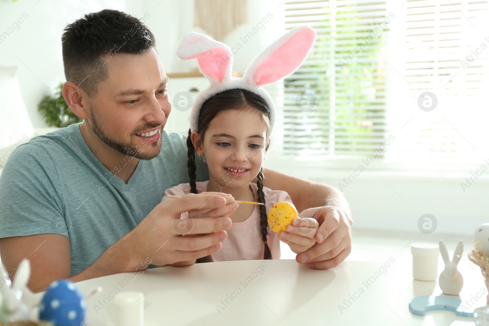 Photo of Happy daughter with bunny ears headband and her father painting Easter egg at home