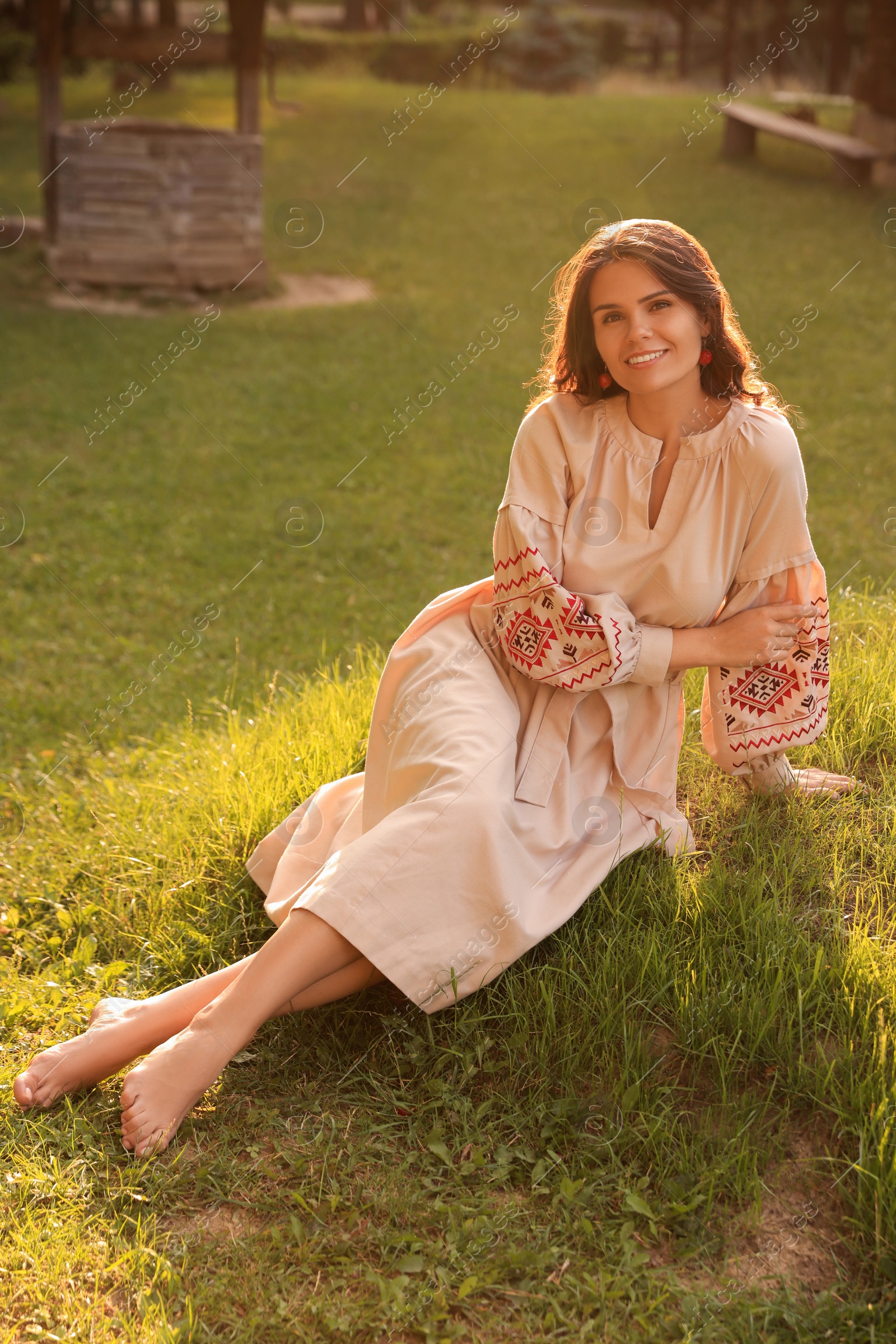 Photo of Beautiful woman in embroidered dress sitting on green grass outdoors. Ukrainian national clothes