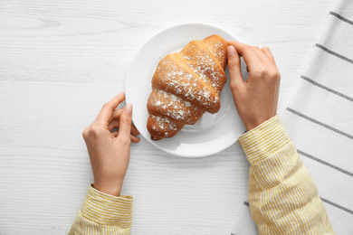 Photo of Woman eating fresh croissant at white wooden table, top view. Delicious morning meal