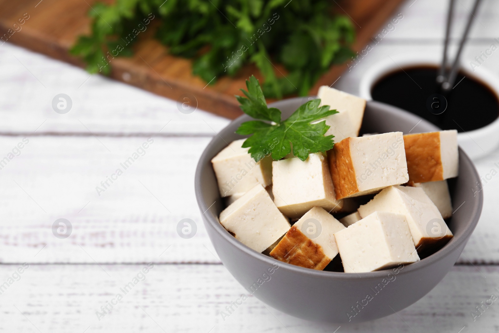 Photo of Bowl of smoked tofu cubes, soy sauce and parsley on white wooden table, closeup. Space for text