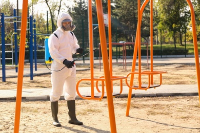 Man in hazmat suit with disinfectant sprayer near swing at children's playground. Surface treatment during coronavirus pandemic