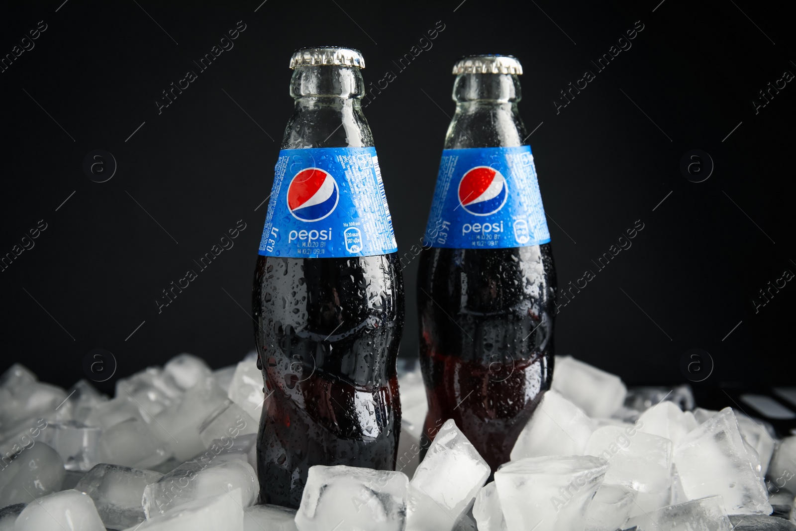 Photo of MYKOLAIV, UKRAINE - FEBRUARY 11, 2021: Glass bottles of Pepsi on ice cubes against black background