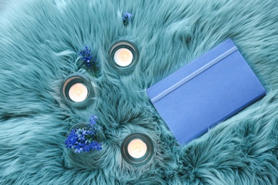 Photo of Burning candles, flowers and notebook on fur fabric, top view