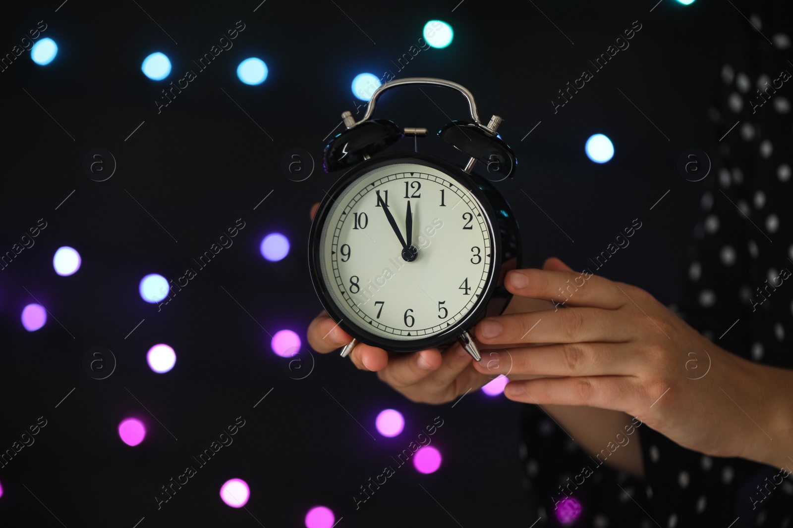 Photo of Woman holding alarm clock against blurred lights, closeup with space for text. New Year countdown