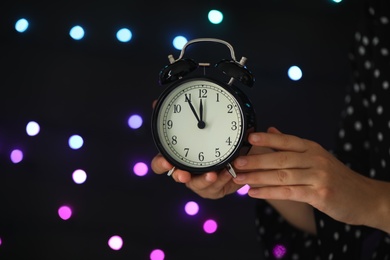 Photo of Woman holding alarm clock against blurred lights, closeup with space for text. New Year countdown