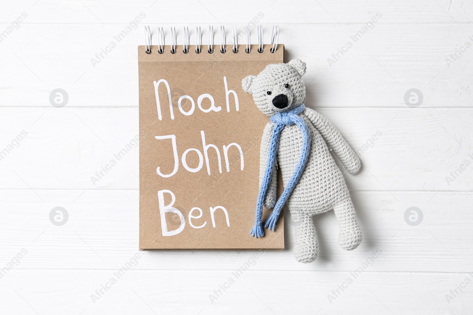 Photo of Notebook with different baby names and toy on white wooden table, flat lay