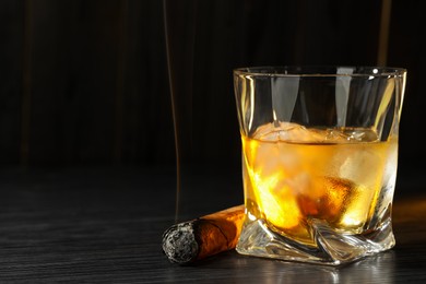 Glass of whiskey with ice cubes and smoldering cigar on black wooden table, closeup. Space for text
