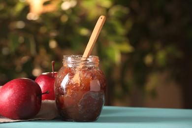 Glass jar with delicious apple jam and fresh fruits on light blue wooden table against blurred background, closeup. Space for text