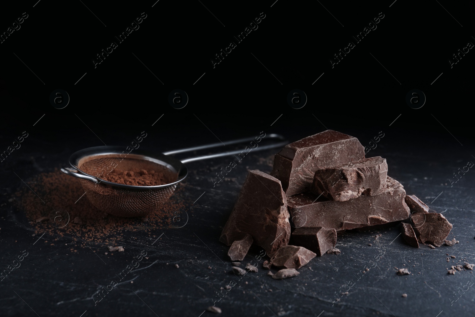 Photo of Pieces of dark chocolate and sieve with cocoa powder on black table