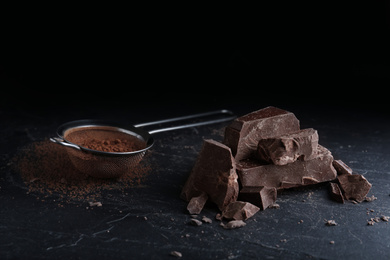 Photo of Pieces of dark chocolate and sieve with cocoa powder on black table