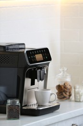 Modern electric coffee machine with cup on white countertop in kitchen