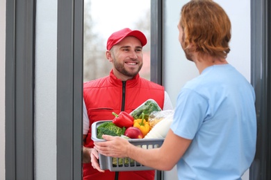 Photo of Courier giving plastic crate with products to customer at home. Food delivery service