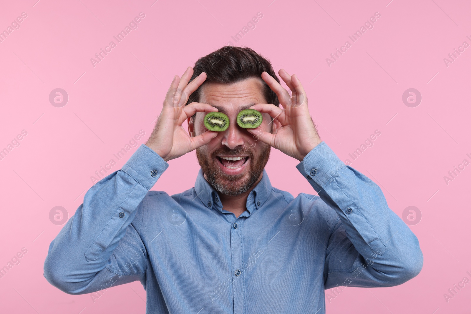 Photo of Man holding halves of kiwi near his eyes on pink background