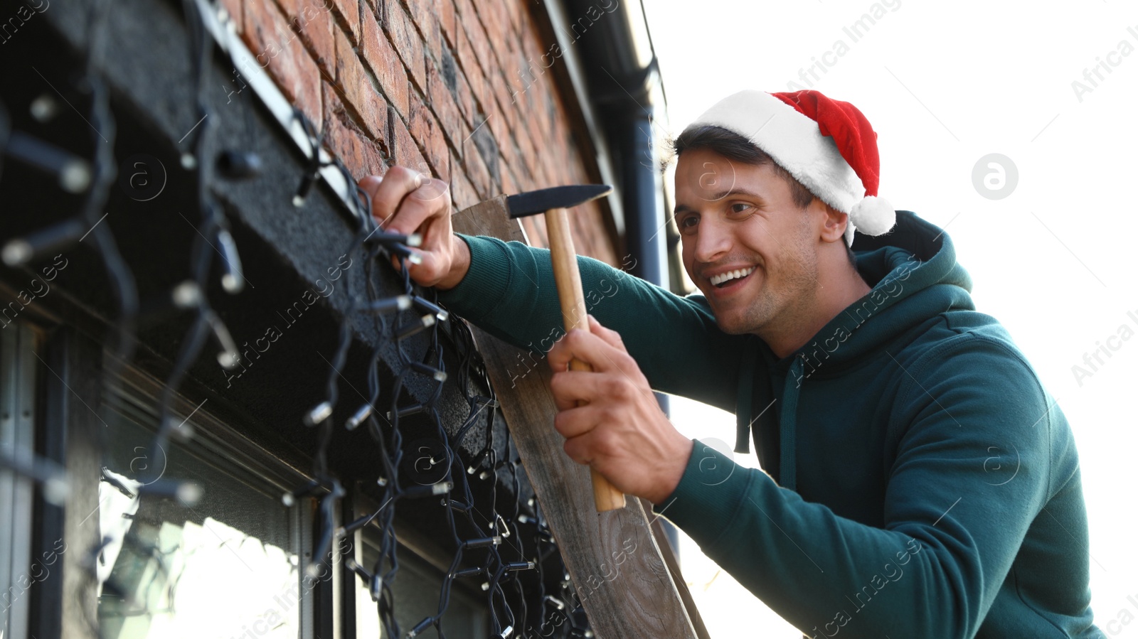 Photo of Man in Santa hat decorating house with Christmas lights outdoors