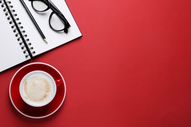 Cup with aromatic cappuccino, notepad, pencil and glasses on red background, flat lay. Space for text